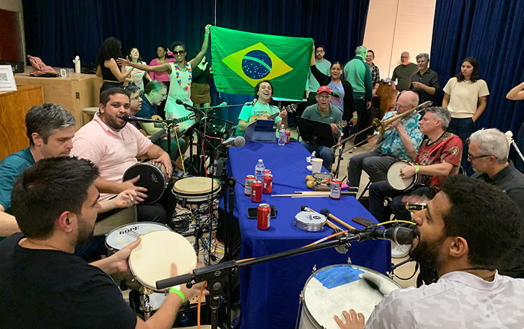 Musicians sit around a table, the flag of Brazil in the background